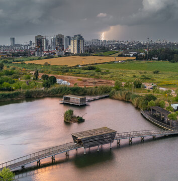 Aerial view of the serene lake reflecting the dramatic sky and distant city skyline under a stormy sky, Hod Hasharon, Center District, Israel.