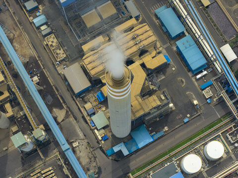 Aerial view of industrial structures and a towering chimney releasing a plume of smoke, contrasting with the geometric layout of buildings, roads, and storage tanks, Coal power plant, Java, Indonesia.