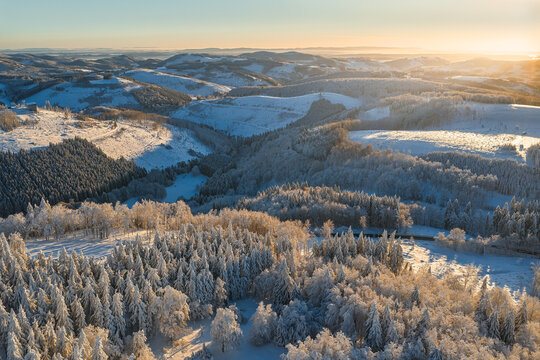 Aerial view of snow-laden forests and rolling hills kissed by the golden light of dawn creating a serene winter wonderland, Winterberg, North Rhine-Westphalia, Germany.