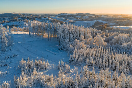 Aerial view of snow-laden trees and hillsides basking in the glow of the morning sun, creating a winter wonderland scene, Winterberg, North Rhine-Westphalia, Germany.