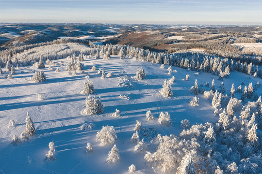 Aerial view of a snowy landscape blanketed in white, trees heavy with snow casting long shadows under a clear blue sky, Winterberg, North Rhine-Westphalia, Germany.