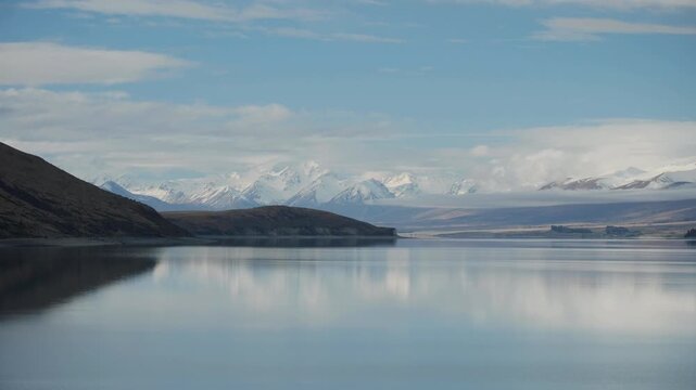 A pastoral and dreamy view of a snow capped mountains and their reflections on a calm water lake at twilight, with some light clouds. a 4K video clip, lake Tekapo, New Zealand.