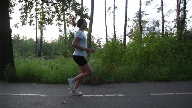 Young woman running on park pathway in slow motion with semi circular tracking camera movement. Shot begins parallel and gradually rotates to follow her motion, creating dynamic cinematic perspective.