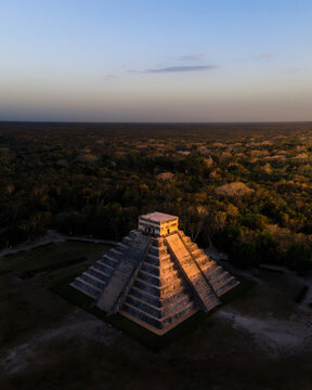 Aerial view of the ancient Mayan pyramid El Castillo bathed in the warm glow of the setting sun, surrounded by a vast, dark forest, Chichen-Itza, Yucatan, Mexico.