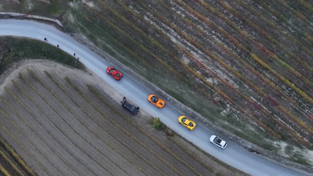 Aerial view of a line of colourful supercars on a winding road cutting through vineyards, La Morra, Piedmont, Italy.
