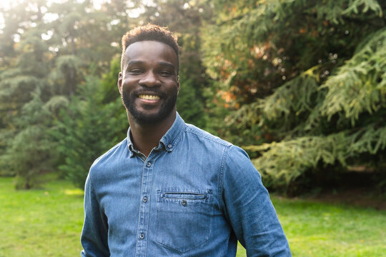 Smiling Man in Blue Shirt Stands in Sunny Park Surrounded by Trees
