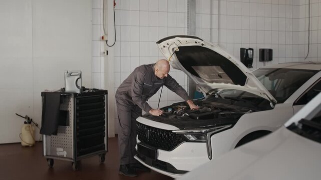 Full shot of mature bald Caucasian male mechanic in gray overalls removing cap from reservoir, then picking up canister from tool cart, then pouring while changing engine oil at garage
