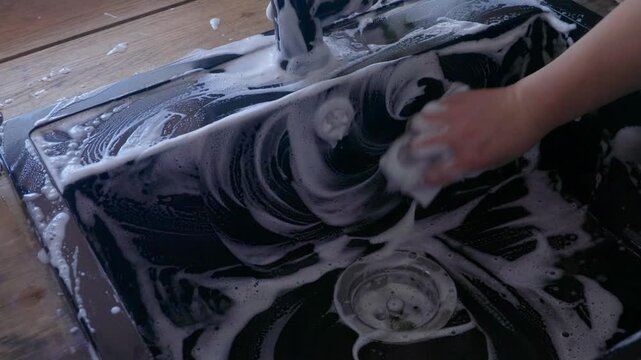 Side view of female hands washing black sink with sponge and soap foam near window. Water running, natural light, everyday domestic cleaning process