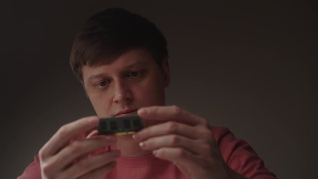 Portrait of focused man inspecting ram module, holding computer memory component between fingers, checking for potential issues during hardware diagnostic process. Concept of computer repairing.
