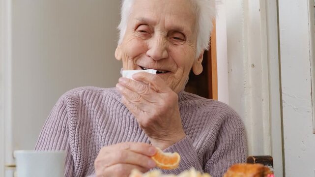 Laughing senior woman enjoys communication with close relatives while sitting at table and putting slice of mandarin or orange into mouth. Dignified aging in family circle. Cheerful elderly people.