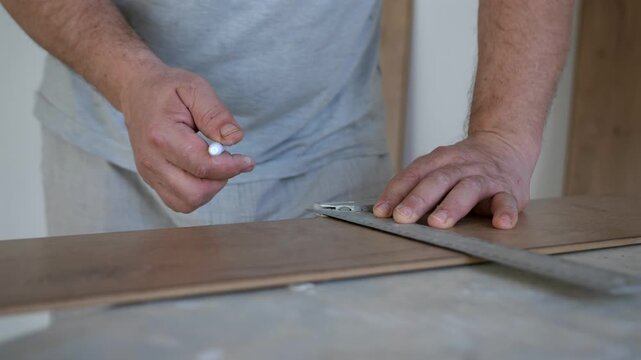 Close up of craftsman measuring and marking laminate flooring panel with tape measure and square ruler before cutting. Home renovation and flooring installation concept.