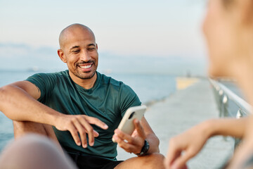 Fototapeta premium Smiling active young couple jogging exercising and having fun and laughing together taking a breakand using a mobile phone after walking and running in the park, fitness and healhty living concept