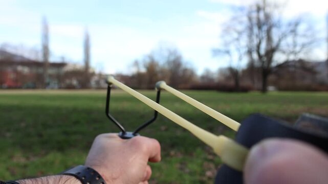 Slow motion close up of hands pulling back a slingshot and releasing a small stone outdoors. Elastic band stretches tightly before the projectile shoots forward in dramatic detail.
