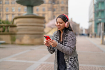 Young woman enjoys music while using a smartphone in a city square near a fountain © Iryna