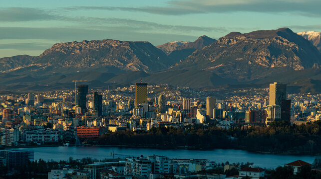 Wide-angle panoramic view of the Tirana city skyline at sunset, featuring the Artificial Lake in the foreground and the snow-capped Dajti Mountains under a soft evening sky.