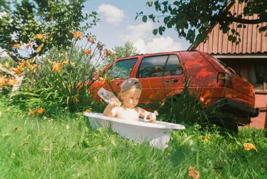 Toddler playing in tub outside near old red car, analog film photo