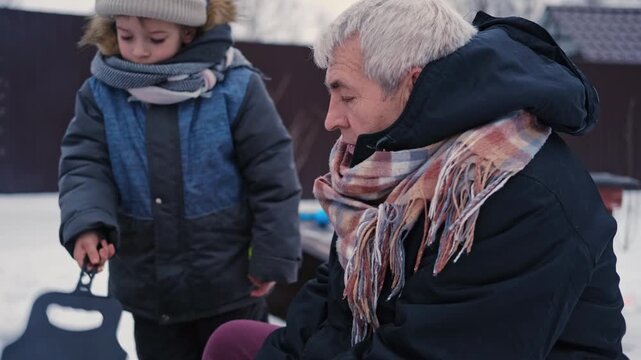 Gray haired grandfather and child by grill roasting sausages on charcoal brazier, bundled in winter coats and scarves on snowy ground. Hands warmed over glowing embers, smoke. Family outdoor barbecue.