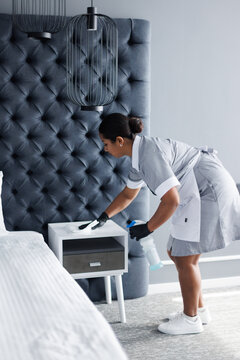 Side view of adult latin maid wiping dust on nightstand in hotel room