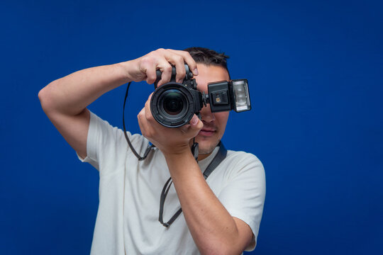 Young man holding professional digital camera with attached flash, taking a picture on a vibrant blue background