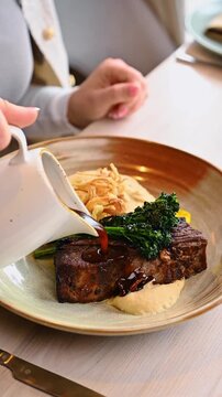 Pouring dark sauce over braised beef short rib. Motion shot of brown gravy being poured from white pitcher onto meat with vegetables.