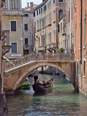 Traditional gondola carrying a couple, being navigated by a gondolier under an arched stone bridge on a narrow residential canal surrounded by historic buildings. © jmag.foto