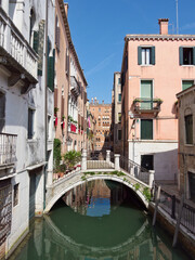 Fototapeta premium Traditional gondola carrying a couple, being navigated by a gondolier under an arched stone bridge on a narrow residential canal surrounded by historic buildings.