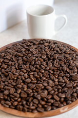 Fototapeta premium Roasted coffee beans in a wooden tray with a white ceramic cup on a light marble table.