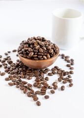 Obraz premium Roasted coffee beans in a wooden bowl with a white cup on a white background, lifestyle morning concept.