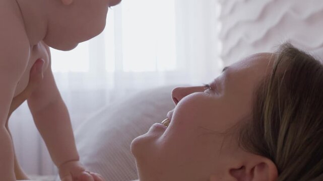 mother lifting baby above chest on bed. morning exercise and play mom with infant with bright window light. cheerful movement and eye contact. happy domestic maternal routine. cozy bonding