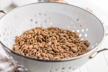 Wholegrain conchiglie pasta in colander on white table.