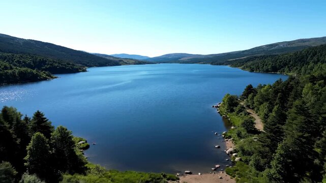 An aerial view captures a serene lake framed by verdant forests under a vast, cloudless sky