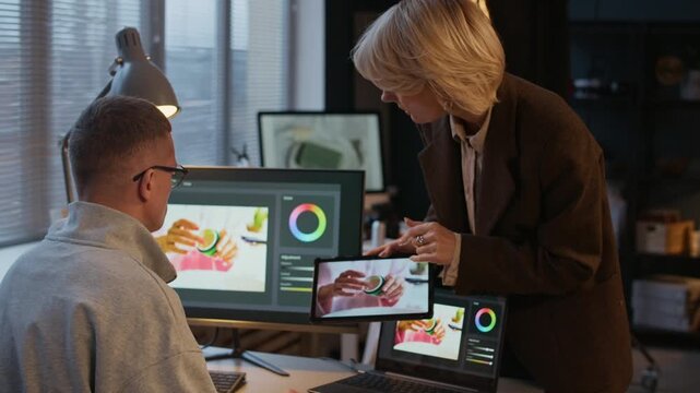 Female producer using tablet, showing image to professional retoucher who sitting in front of her, listening to her attentively and looking at display of tablet