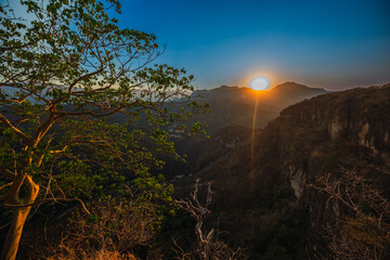 Beautiful landscape of a ravine between mountains, a small valley and many trees, in the foreground appears a huge yellow fig tree and in the background the sun is setting.
