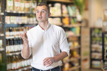Fototapeta premium Positive young man walking through organic store aisle, examining with interest and focus wide range of products on shelves
