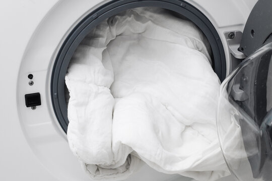 White bedding inside washing machine drum. Laundry cycle with sheets and duvet being washed. Household textile cleaning and hygiene care. Fresh linens prepared through domestic washing process.