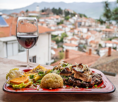 A delicious Balkan dinner platter with grilled meat, rice, polenta, and salad, paired with a glass of red wine, all overlooking a blurred Ohrid city panorama.