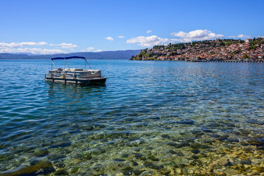 Undisturbed, blue waters of Lake Ohrid reflect the bright atmosphere as a pontoon boat rests gently. In the distance, the charming, red-roofed town of Ohrid ascends a verdant hill.