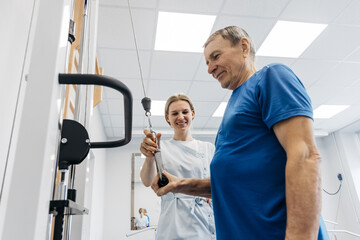 Female physical therapist assists elderly male patient with therapeutic exercise using cable machine in bright rehabilitation center with modern equipment