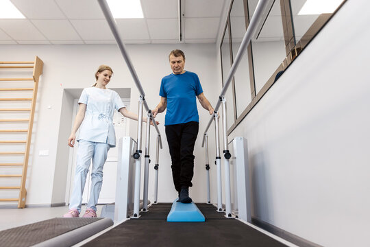 Male patient performing leg rehabilitation exercises on a balance platform with assistance from a female physical therapist in a modern therapy room