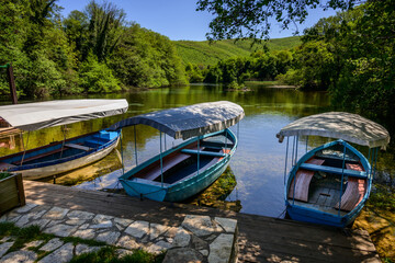 Three rowboats, adorned with protective awnings, moored by a rustic wooden dock on the pure waters of a still forest river, surrounded by lush greenery near Saint Naum Monastery in North Macedonia..