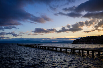 A long pier extends into the choppy, dark waters of Lake Ohrid under a dramatic twilight sky,...