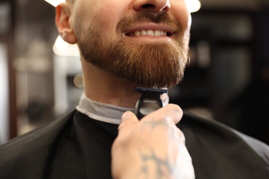Stylist trimming man's beard with hair clipper in barbershop, closeup
