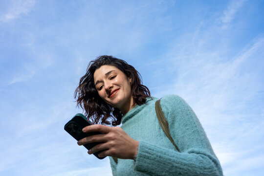 Woman Smiling and Using Smartphone Against Blue Sky Backdrop