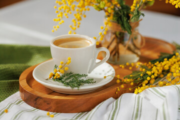A cup of coffee placed on a wooden tray with vibrant mimosa flowers, creating a bright and peaceful spring ambiance.