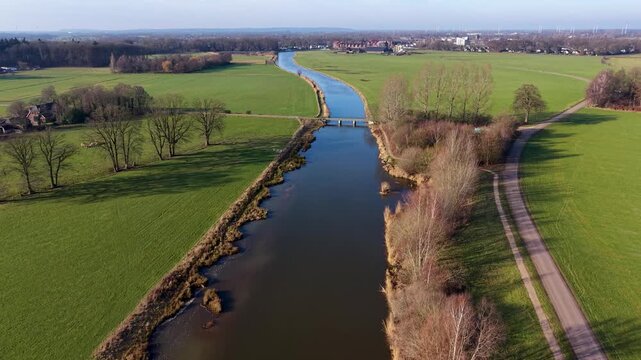 Aerial view of a rural landscape with a canal curving through green fields, two small road bridges crossing it, and a nearby house with outbuildings set among open farmland.