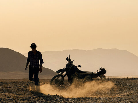 Man Walks Away From Motorcycle in a Dusty Desert Landscape at Sunset
