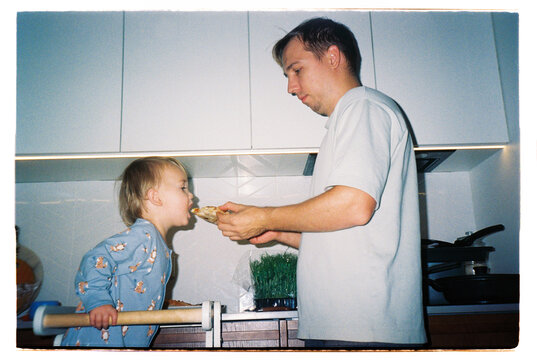 Father feeding toddler a slice of pizza in the kitchen, film photo