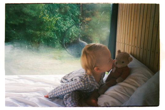 Toddler in pajamas kissing teddy bear on bed by cabin window - film 