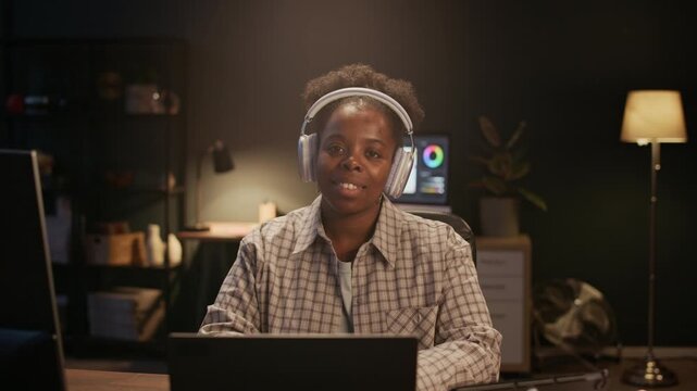 Medium shot of female African American retoucher enjoying listening to music via headphones while working on computer and looking at camera