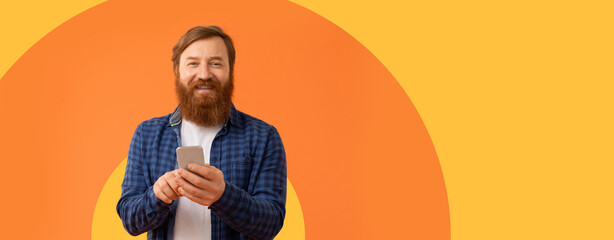 A man stands in front of a bright orange background. He holds a smartphone in his hands and smiles....